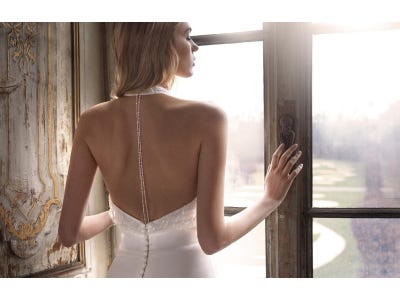 Modella di spalle che si affaccia a una finestra e indossa un abito da sposa con schiena scoperta e dettagli in gioiello.