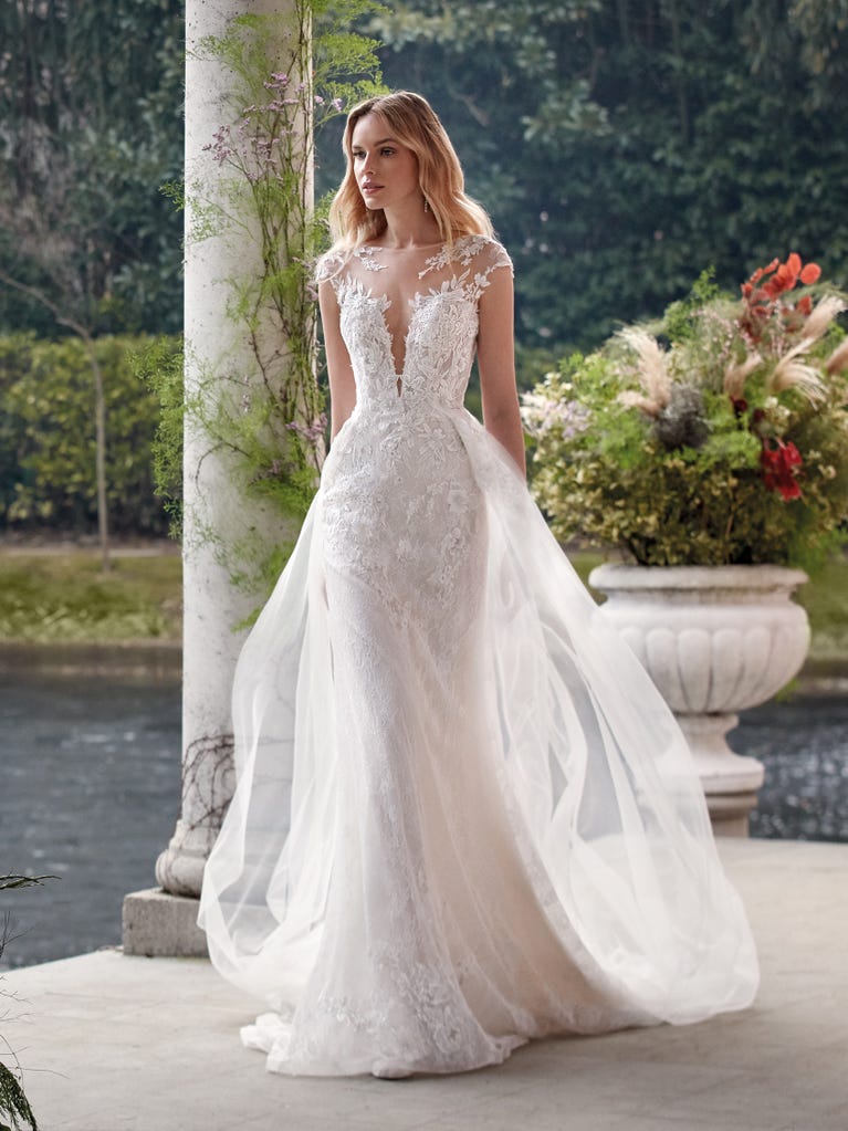 Flared wedding dress with crepe skirt and a tulle cape embroidered with silver beads. The three-dimensional flowers and minimalist, geometric superimpositions of fabrics create a contemporary, unique look for brides who want to surprise.