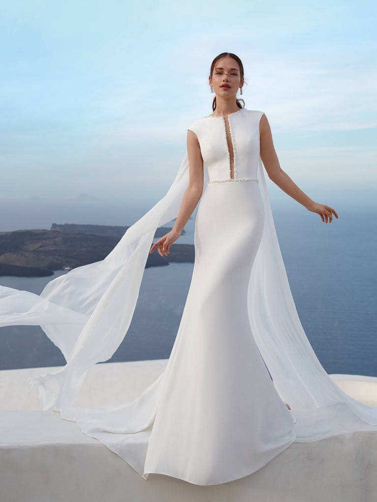 Flared wedding dress with crepe skirt and a tulle cape embroidered with silver beads. The three-dimensional flowers and minimalist, geometric superimpositions of fabrics create a contemporary, unique look for brides who want to surprise.