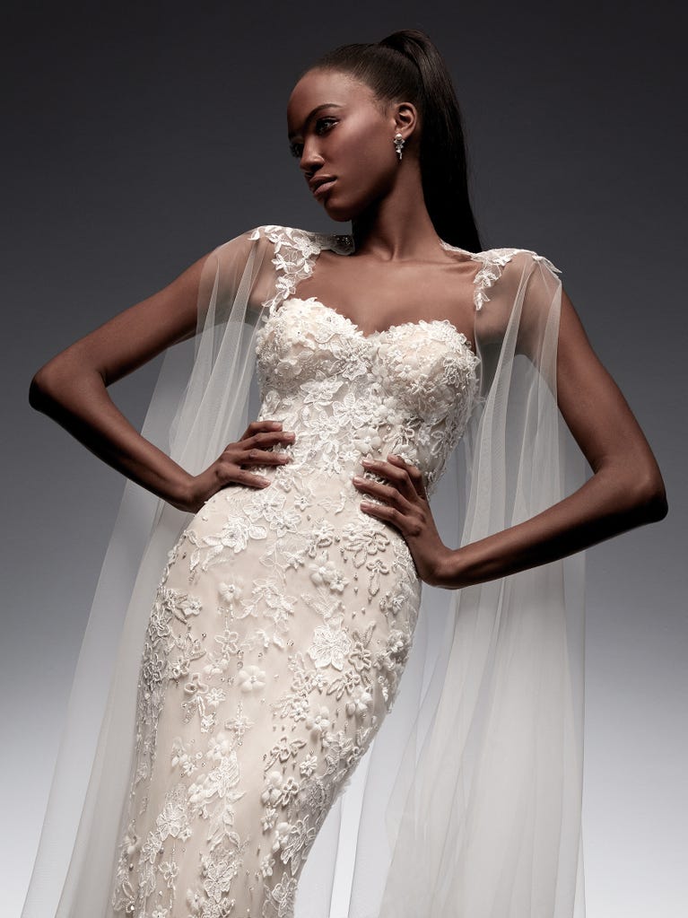 Flared wedding dress with crepe skirt and a tulle cape embroidered with silver beads. The three-dimensional flowers and minimalist, geometric superimpositions of fabrics create a contemporary, unique look for brides who want to surprise.
