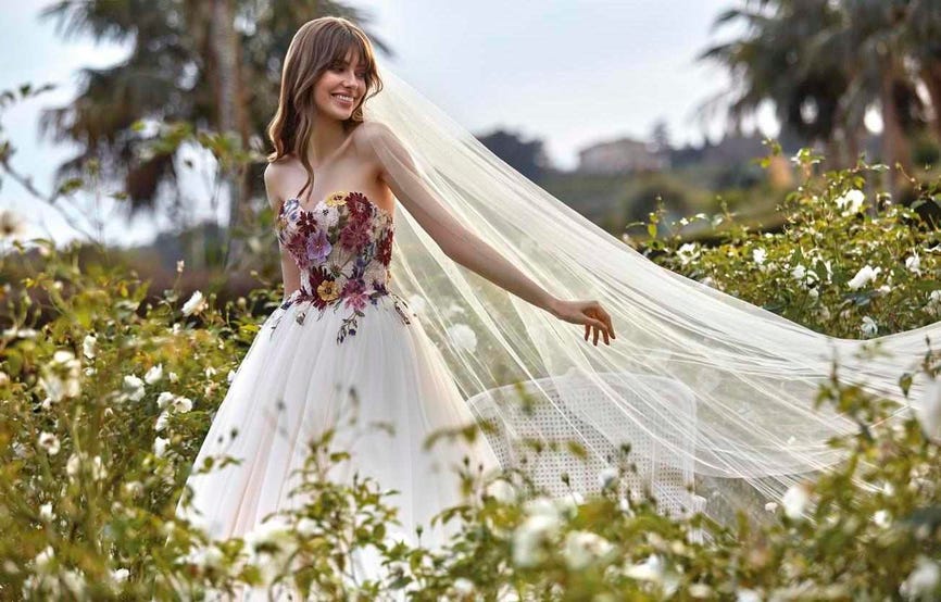 Sposa in un giardino con tanti fiori e con un vestito a principessa con corpetto floreale e colorato e gonna in tulle.