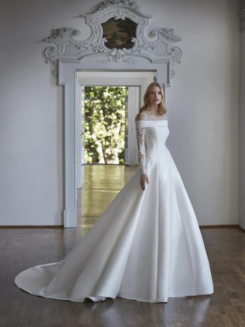 Jeune femme vêtue d'une robe de mariée col bateau, se tenant dos à une porte menant vers un extérieur naturel et fleuri Robe de mariée classique