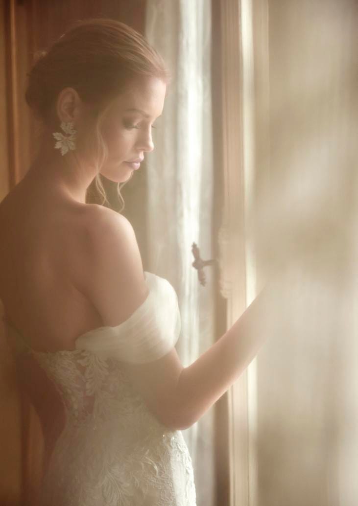 Sposa di profilo che indossa un vestito con spalline cadenti in tulle e ha un acconciatura raccolta e ciuffi pendenti.