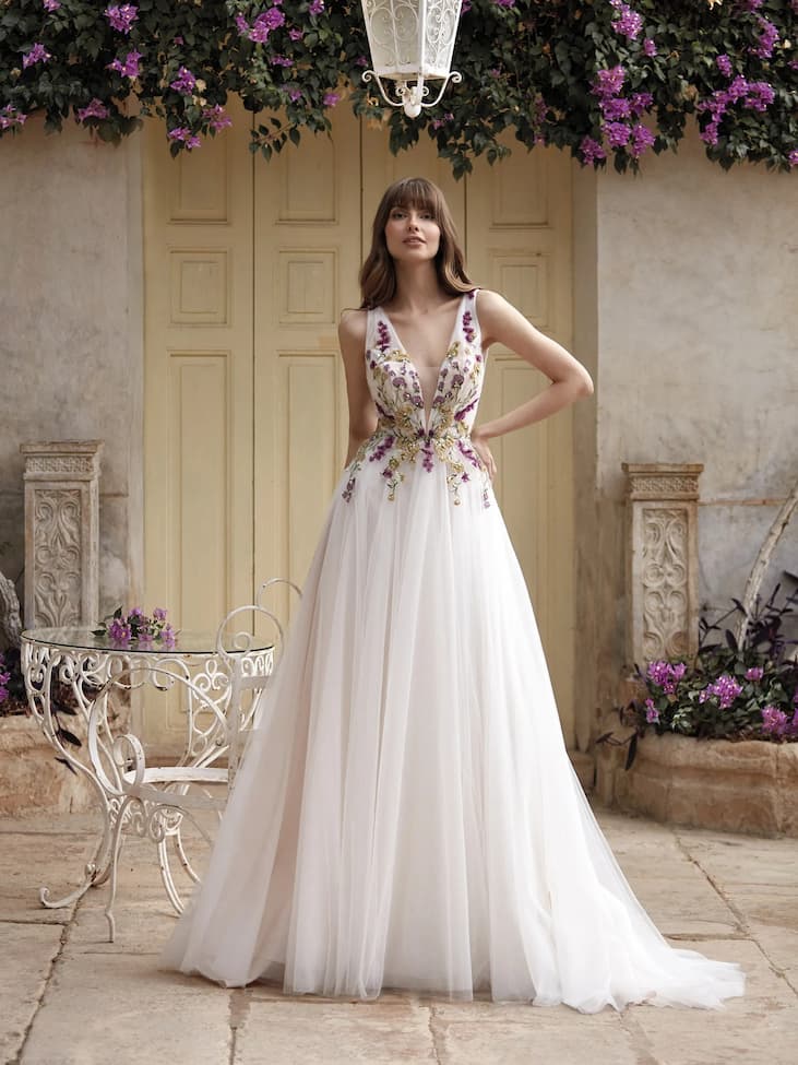 Robe de mariée rose clair à fleurs Une mariée se tenant au milieu d'un patio fleuri, devant une table où elle a déposé son bouquet mauve