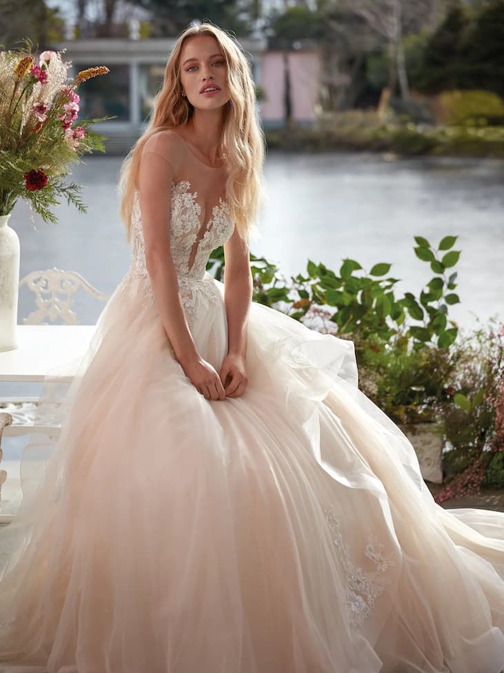 Une jeune mariée assise au bord d’un lac, vêtue d’une robe de mariée rose tendre entourée de magnifiques fleurs.