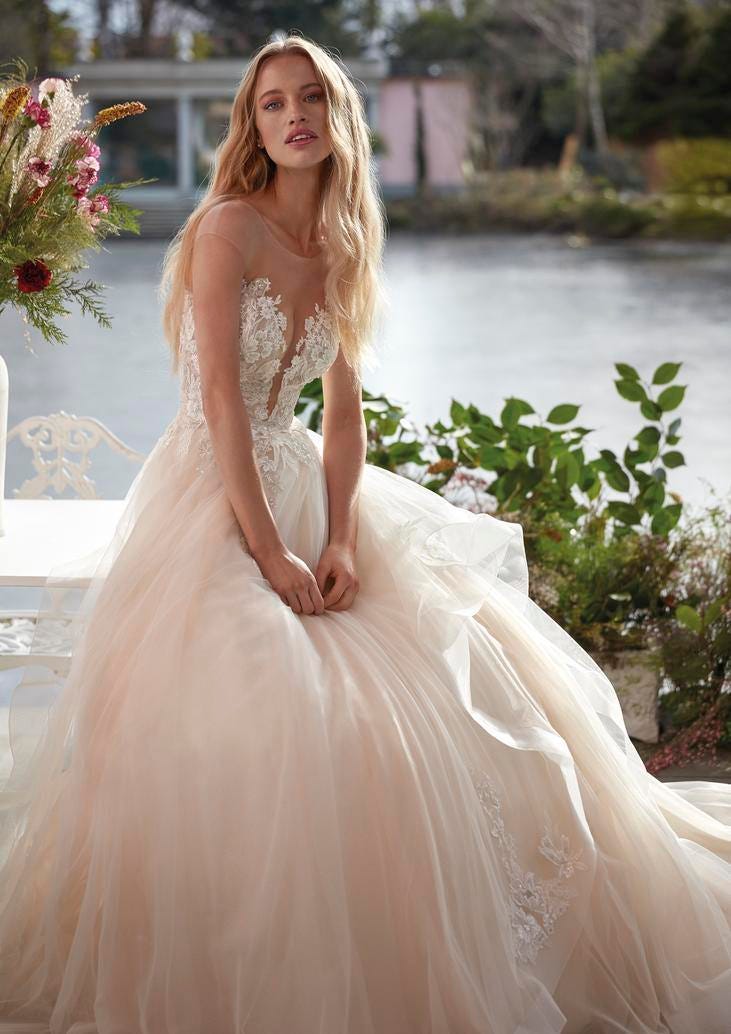 Une femme vêtue d'une robe de mariée en tulle rose sans manches. Le tulle crée un léger volume sur la jupe ornée de broderies